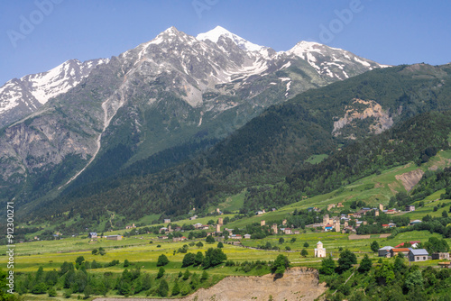A village in a valley below a mountain. The church and defensive towers are visible. Snow capped mountains in the background