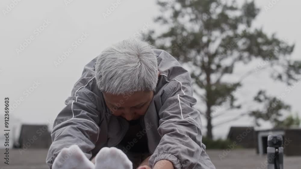 Slowmo shot of senior Asian woman doing stretching exercise on yoga mat in open air Slowmo shot of senior Asian woman doing stretching exercise on yoga mat in open air