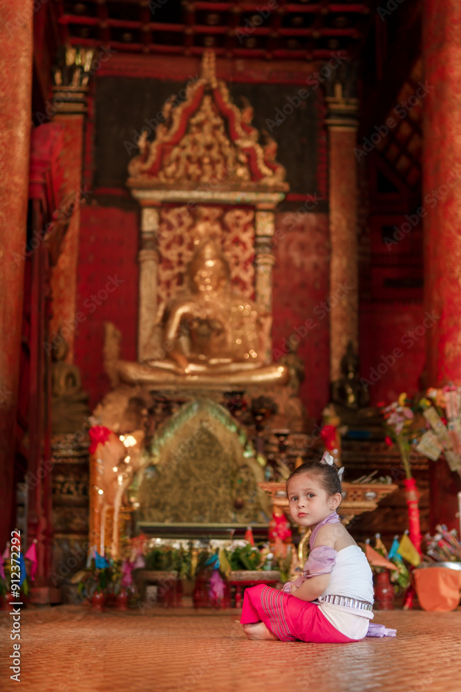 Obraz premium A little girl sits in front of a statue of Buddha. The statue is surrounded by flowers and candles. The girl is wearing a white dress and a pink headband. The scene is peaceful and serene
