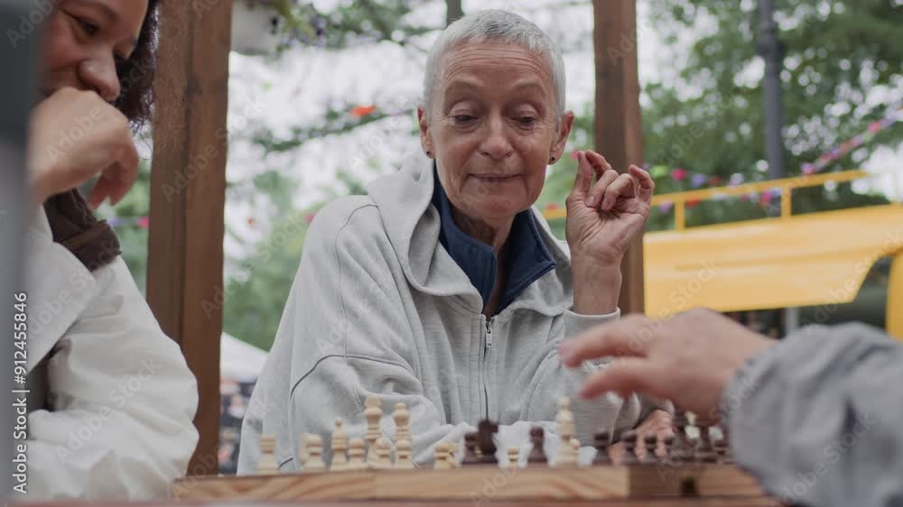 Waist up of joyful Caucasian senior woman with short grey hair having fun while playing chess with diverse female friends at wooden table in park on cloudy day Waist up of joyful Caucasian senior woman with short grey hair having fun while playing chess with diverse female friends at wooden table in park on cloudy day