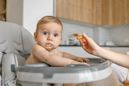 mother hand feeding a baby from a spoon. Mom giving a 9 month baby porridge. Meal time. Little boy eats sitting on baby's feeding chair.