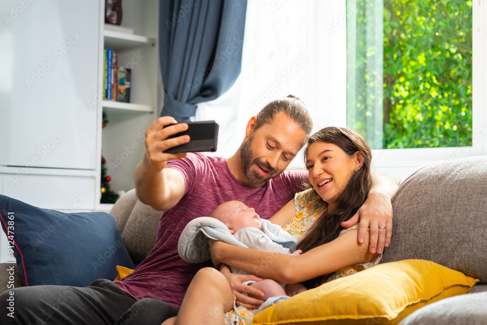 Happy Caucasian couple mother and father using mobile taking selfie together with newborn baby son on sofa in living room. Parents spending time with infant child boy at home. Family bonding concept. Happy Caucasian couple mother and father using mobile taking selfie together with newborn baby son on sofa in living room. Parents spending time with infant child boy at home. Family bonding concept.