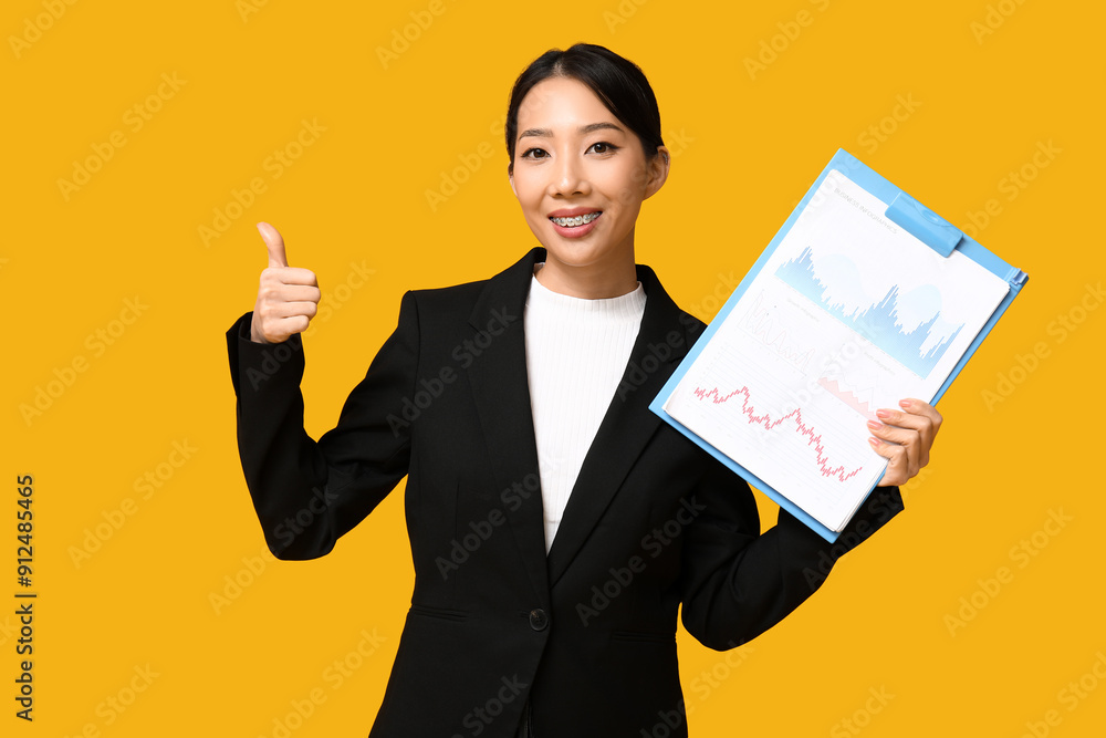 Portrait of young Asian businesswoman with charts showing thumb-up on yellow background Portrait of young Asian businesswoman with charts showing thumb-up on yellow background
