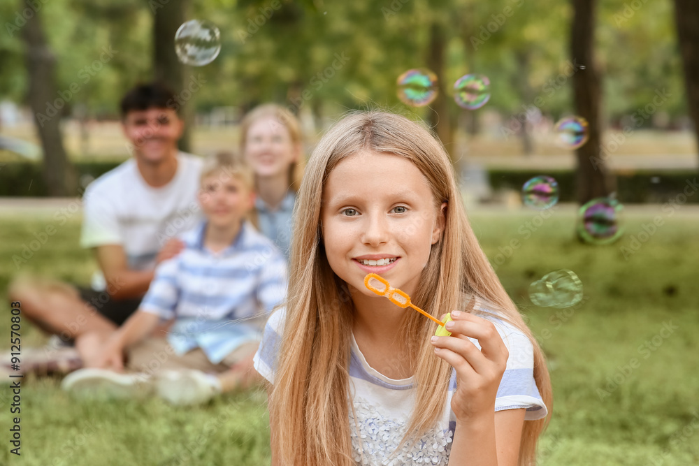 Happy family with cute daughter blowing soap bubbles while sitting on grass in park Happy family with cute daughter blowing soap bubbles while sitting on grass in park
