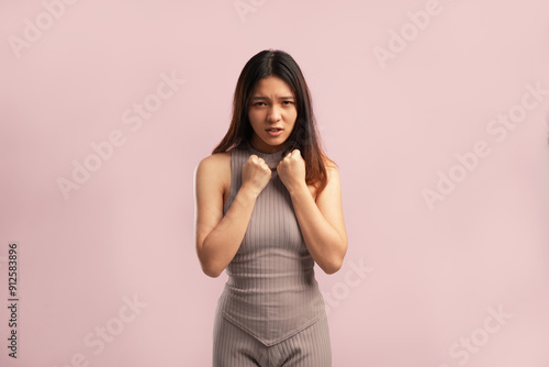 A woman with long hair is standing in front of a pink background. She is wearing a gray shirt and has her arms crossed. Concept of frustration or annoyance, as the woman is in a bad mood