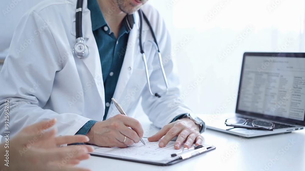 Doctor and a patient. The physician, wearing a white medical coat over a green shirt, is filling out a medical record form during a consultation in the clinic. Medical service Doctor and a patient. The physician, wearing a white medical coat over a green shirt, is filling out a medical record form during a consultation in the clinic. Medical service