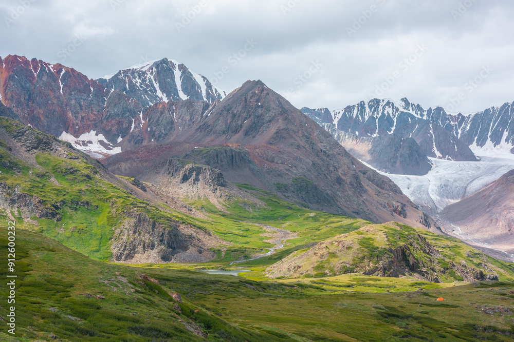 Vivid dramatic alpine scenery with green hills and rocks with view to high rocky pointy peak, sharp rockies, large snow-capped mountain top, snowy range and big glacier tongue under gray cloudy sky. Vivid dramatic alpine scenery with green hills and rocks with view to high rocky pointy peak, sharp rockies, large snow-capped mountain top, snowy range and big glacier tongue under gray cloudy sky.