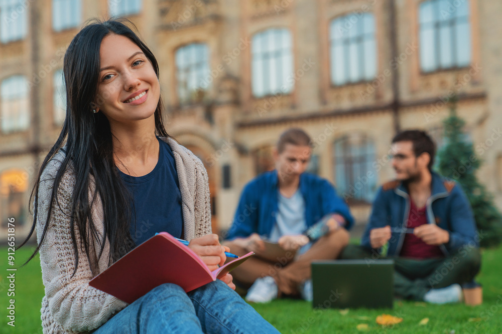 Portrait of young female student woman girl noting doing homework task sitting on the grass while two males college students classmates boys using laptop in background at university campus lawn Portrait of young female student woman girl noting doing homework task sitting on the grass while two males college students classmates boys using laptop in background at university campus lawn
