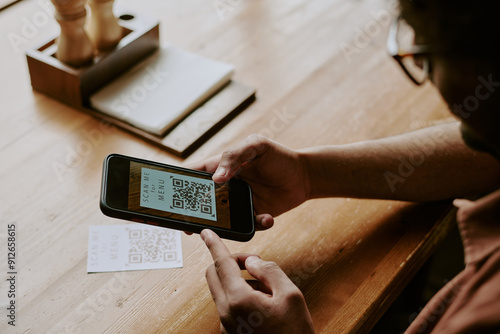 Close-up of hands scanning QR code with smartphone on wooden table, device visible in background displaying QR code, reflection of glasses seen in user's hand photo