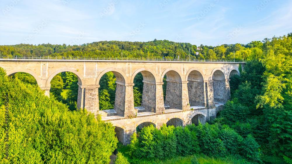 Fototapeta premium The Sychrov Railway Bridge stands majestically with graceful arches, surrounded by lush greenery. The bright sky enhances the tranquil setting.