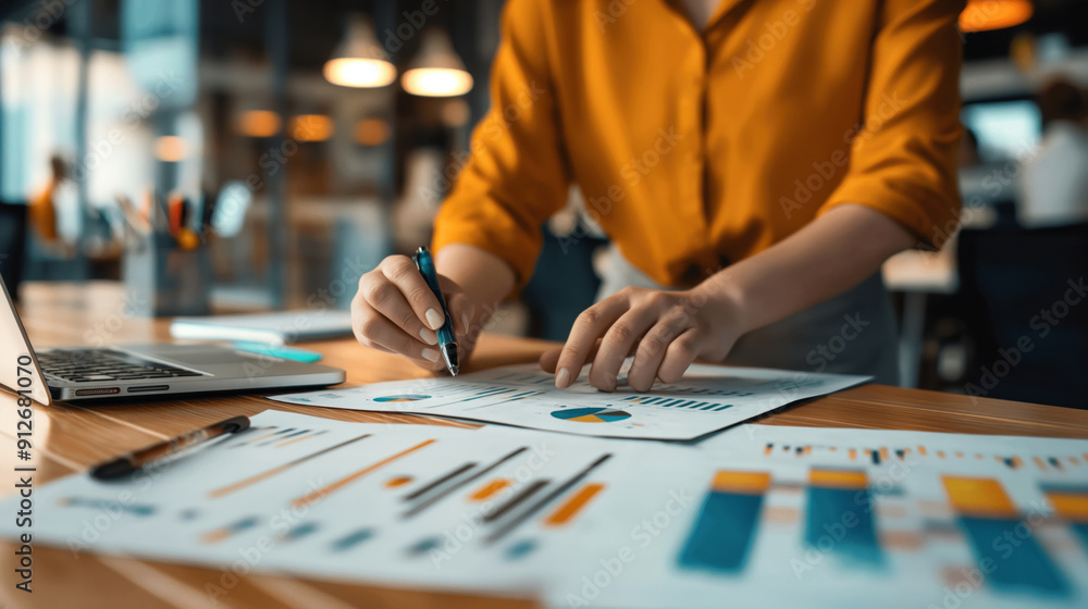 A person in a bright shirt is analyzing graphs and charts on a desk in an office environment. A laptop is open nearby with blurred background elements. A person in a bright shirt is analyzing graphs and charts on a desk in an office environment. A laptop is open nearby with blurred background elements.