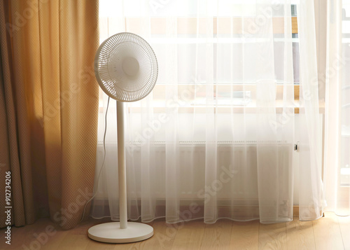 A white, modern pedestal fan is positioned near a window with curtains. The fan's design is sleek and minimalistic, complementing the room's cozy and serene decor.