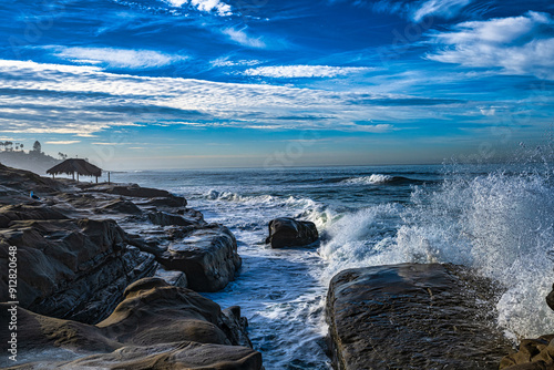 2023-12-31 ROCKY SHORELINE AT THE WINDANSEA BEACH WITH A GRASS HUT AND WAVES CRASHING ON THE ROCKS SPRAYING WATER IN LA JOLLA CALIFORNIA NEAR SAN DIEGO photo