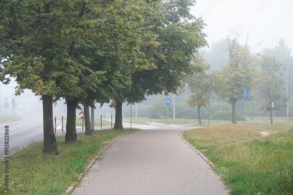 Morning fog in city. Sidewalk and trees alongside a bike path. The city awakens to life in the early hours. Serene urban landscape with misty surroundings. Mystic horror scenery. Scaryy landscape. Morning fog in city. Sidewalk and trees alongside a bike path. The city awakens to life in the early hours. Serene urban landscape with misty surroundings. Mystic horror scenery. Scaryy landscape.
