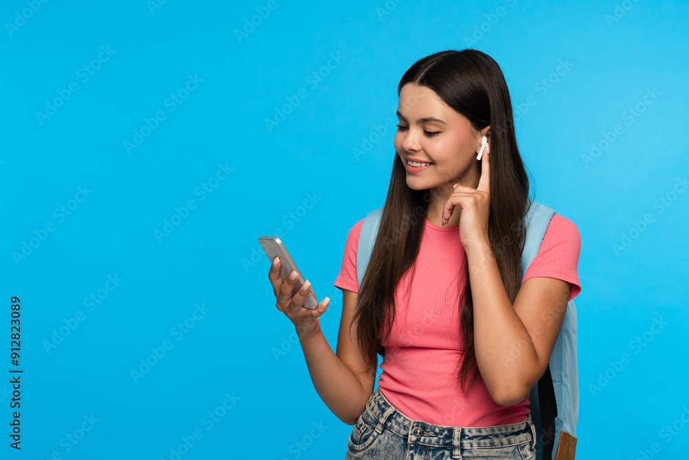 Cheerful latin hispanic girl high school female pupil student teenager with bag holding smartphone wearing earbud headphones using wireless earphones isolated on blue background copy space Cheerful latin hispanic girl high school female pupil student teenager with bag holding smartphone wearing earbud headphones using wireless earphones isolated on blue background copy space