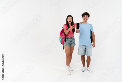 Full length of high school pupils students teenagers classmates boy girl friends with bags holding using smart mobile phone showing mock up screen pointing fingers isolated on white background.