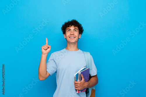 Foto Smiling boy guy high school male pupil student teenager with bag holding books copybooks having idea pointing index finger up isolated on blue background