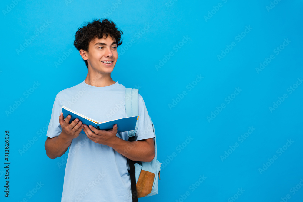 Smiling boy guy high school male pupil student teenager with bag holding open books copybooks isolated on blue background looking empty copy space for advertisement. Education back to school concept Smiling boy guy high school male pupil student teenager with bag holding open books copybooks isolated on blue background looking empty copy space for advertisement. Education back to school concept
