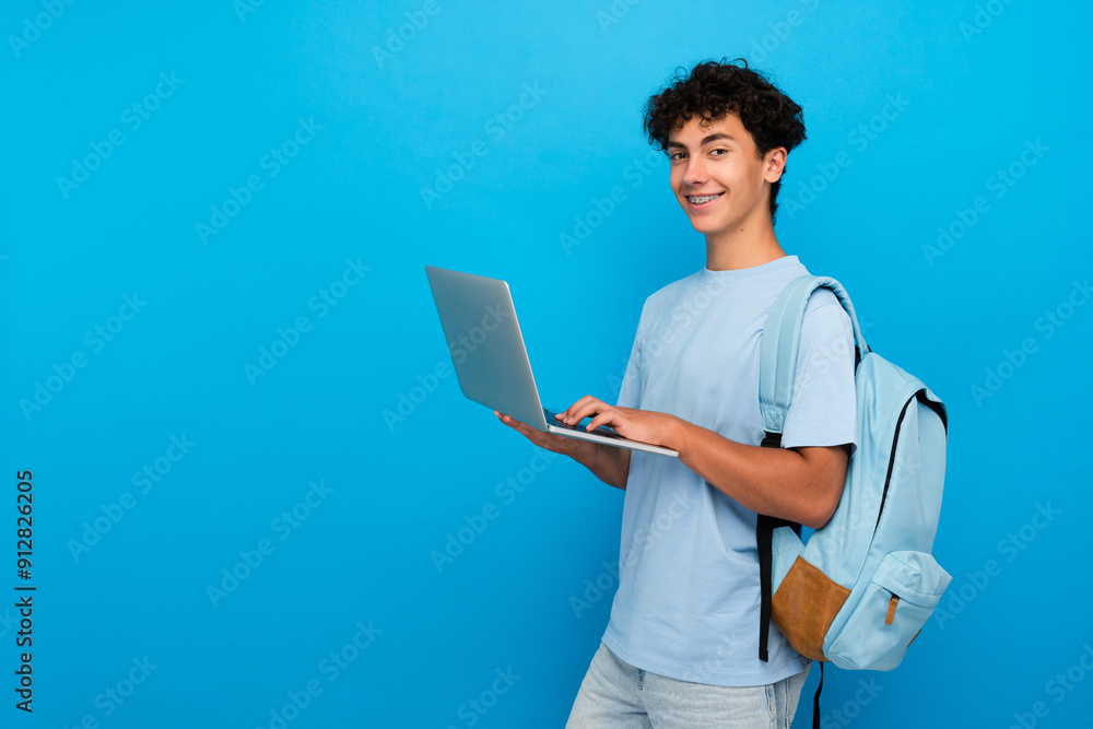 Smiling boy guy high school male pupil student teenager with backpack holding using laptop computer for distant studding ,e-learning doing homework online isolated in blue background copy space Smiling boy guy high school male pupil student teenager with backpack holding using laptop computer for distant studding ,e-learning doing homework online isolated in blue background copy space