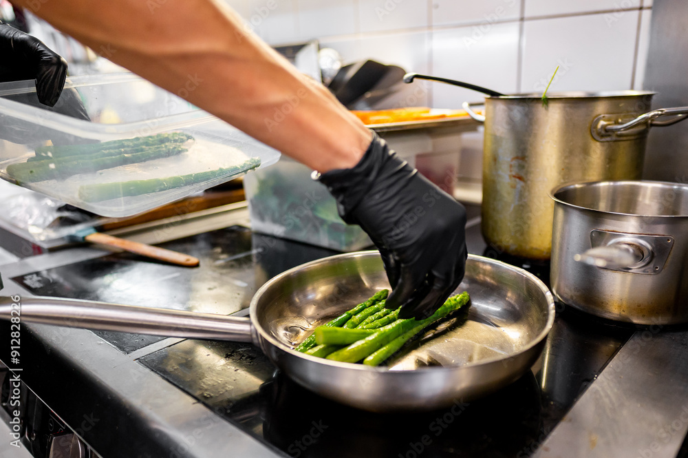 Φωτογραφία Close-up of a hand in a black glove cooking fresh asparagus in a stainless steel pan on a gas stove, surrounded by kitchen utensils and pots