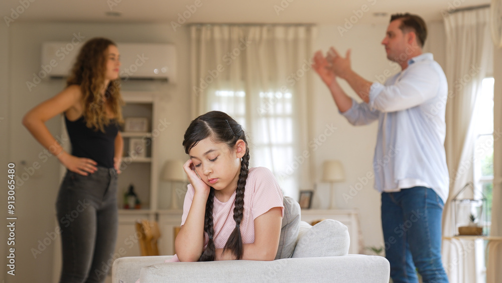 Annoyed and unhappy young girl sitting on sofa trapped in middle of tension by her parent argument in living room. Unhealthy domestic lifestyle and traumatic childhood develop to depression Synchronos Annoyed and unhappy young girl sitting on sofa trapped in middle of tension by her parent argument in living room. Unhealthy domestic lifestyle and traumatic childhood develop to depression Synchronos