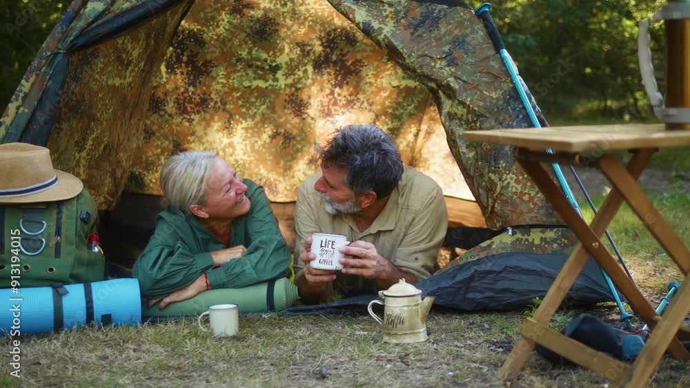 Smiling senior couple lying in tent and enjoying morning coffee while camping in nature Smiling senior couple lying in tent and enjoying morning coffee while camping in nature