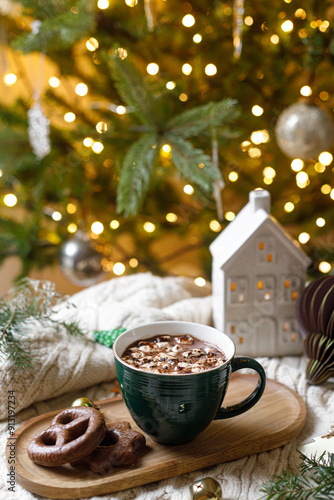 Warm cup of cocoa with marshmallows and chocolate cookies, christmas ornaments, cozy knitted sweater and fir branches on background of stylish christmas tree with golden lights. Winter mood photo