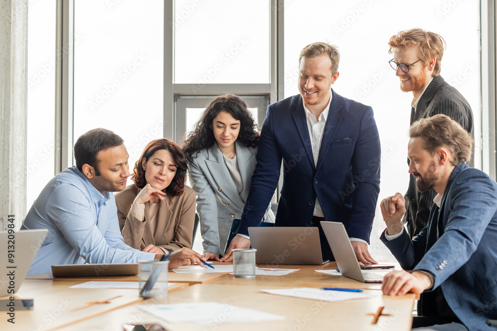 A group of diverse business professionals are gathered around a table in a modern office, working together on a project. They are looking at a laptop screen and discussing ideas A group of diverse business professionals are gathered around a table in a modern office, working together on a project. They are looking at a laptop screen and discussing ideas