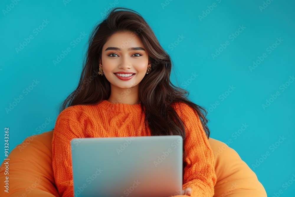 Smiling young woman in orange sweater sits on a beanbag chair and uses a laptop. Smiling young woman in orange sweater sits on a beanbag chair and uses a laptop.