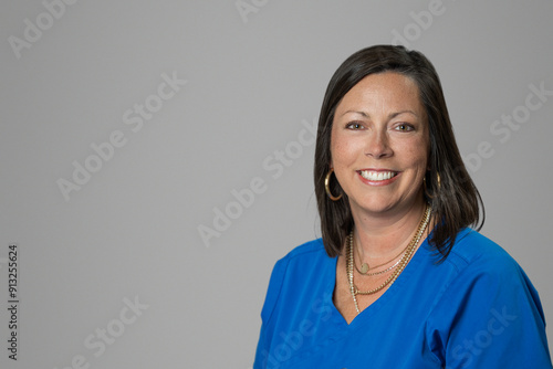 Smiling headshot of Caucasian brunette middle age medical worker woman in a blue scrubs top in a studio on gray grey backdrop