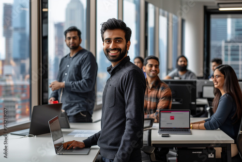 An office with Indian business people, a young businessman standing happily looking at the camera.