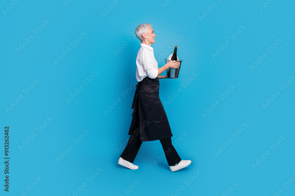 Full length photo of pensioner lady carry walk profile hold bucket wine restaurant dressed waitress uniform isolated on blue background Full length photo of pensioner lady carry walk profile hold bucket wine restaurant dressed waitress uniform isolated on blue background