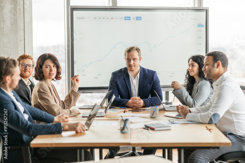 A group of business professionals are gathered around a table in a modern office, discussing a growth chart displayed on a large screen. The team is engaged in a lively conversation photo