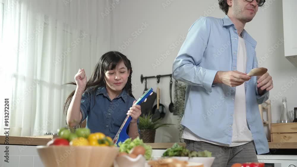 Caucasian father dancing while asian girl playing ukulele at kitchen. Energetic dad and cute daughter spend time together and moving to music in lively mood surrounded with vegetable placed. Pedagogy. Caucasian father dancing while asian girl playing ukulele at kitchen. Energetic dad and cute daughter spend time together and moving to music in lively mood surrounded with vegetable placed. Pedagogy.