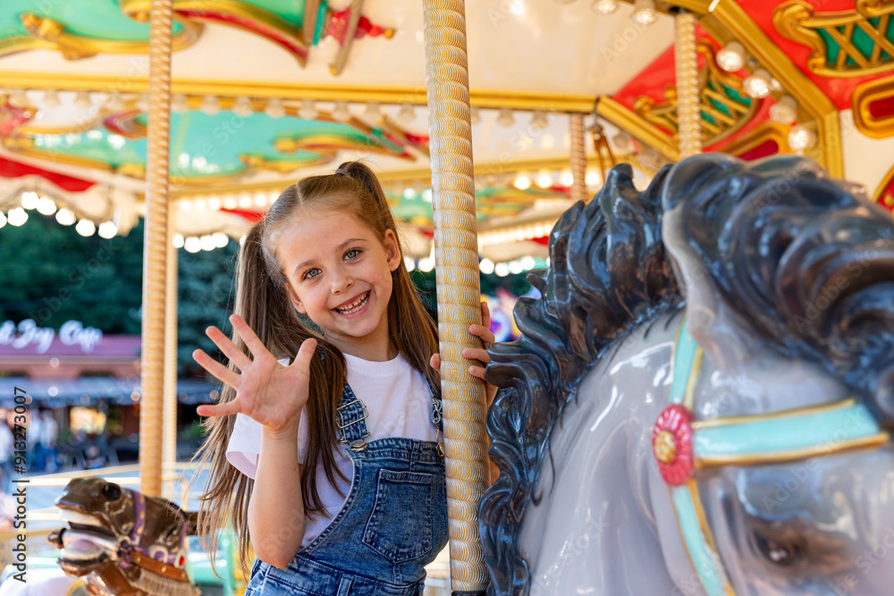 Fototapeta premium Happy emotional child riding on a merry go round at amusement park. Little girl playing on carousel, summer fun, happy childhood, vacation and entertainment concept