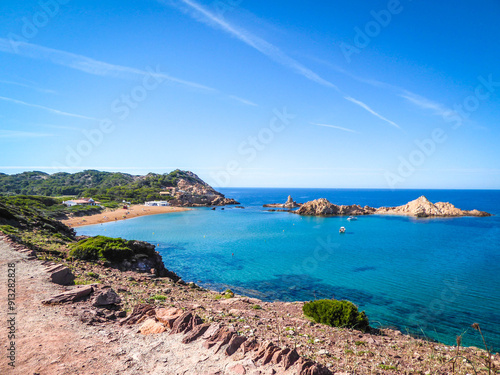 Idyllic panoramic view of Cala Pregonda in Menorca: a circular beach with reddish sand, clear blue ocean, and rocky outcrops on the horizon. A postcard-perfect scene on a clear day