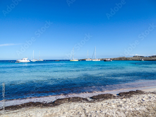 Binibeca Beach in Menorca: clear blue water, white sailboats on the horizon, and seaweed on the shore. Tranquil and picturesque coastal scene
