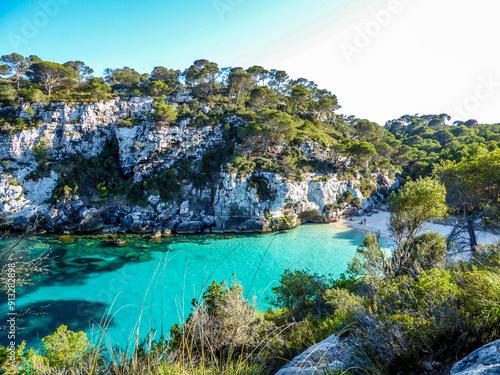 Aerial view of Cala Turqueta in Menorca: crystal-clear turquoise sea, white sand, and green pines under a clear sky. Natural and idyllic Mediterranean landscape.