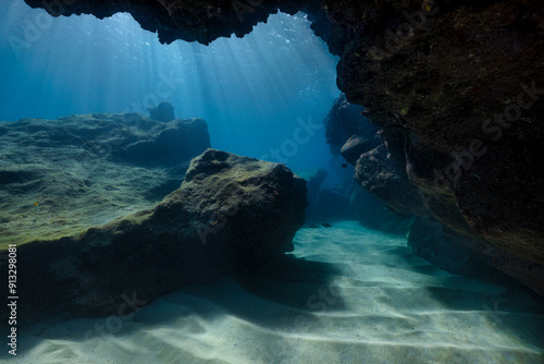 An underwater scenic view from inside a cave as the sun penetrates the water, raining sun rays down on the sandy and rocky sea floor