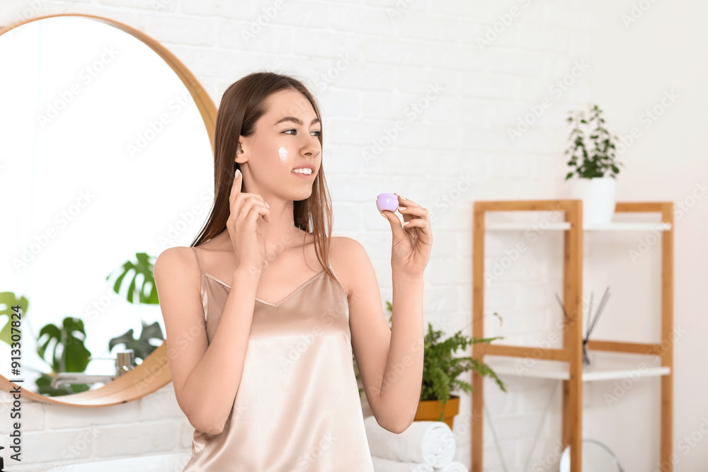 Beautiful young woman with jar applying cream on face in bathroom Beautiful young woman with jar applying cream on face in bathroom
