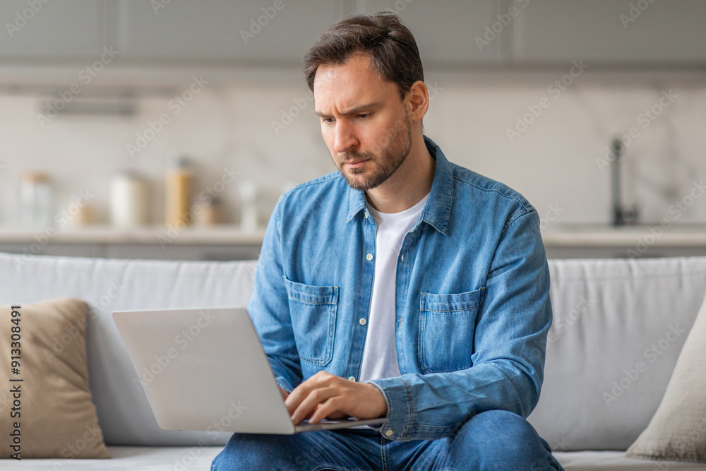 A man is sitting on a white couch in a living room, working on a laptop. He is wearing a blue button-down shirt and blue jeans. He looks focused on his computer A man is sitting on a white couch in a living room, working on a laptop. He is wearing a blue button-down shirt and blue jeans. He looks focused on his computer