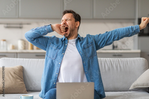 A man wearing a blue shirt is sitting on a couch with a laptop in front of him. He is yawning with his mouth wide open and his eyes closed. His right arm is stretched out in front of him.