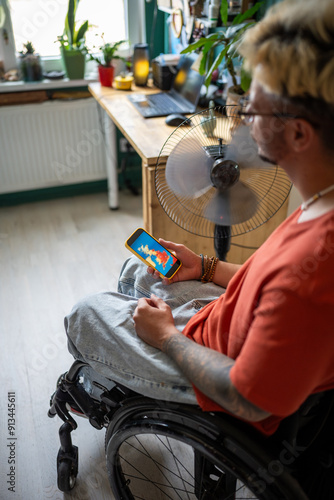 Disabled man in wheelchair looks at weather forecast on smartphone screen, sits by electric fan at home during the extreme summer heat wave, suffers from stuffiness and lack of air conditioning. photo