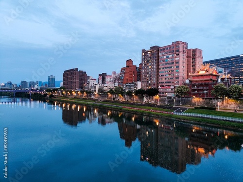 A tranquil waterfront cityscape at dusk with illuminated buildings reflecting in the calm river. The skyline showcases a blend of modern and traditional architecture under a serene evening sky. photo