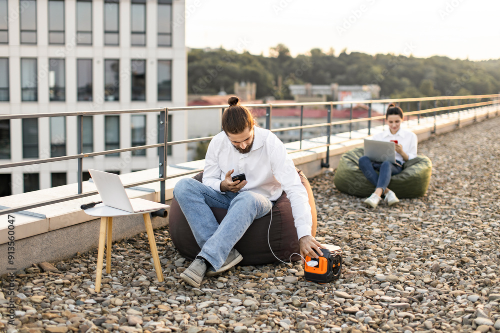 Businessman sitting on beanbag chair and plugs phone charging cable into portable power station on roof of house. Colleague in background working on laptop, creating modern rooftop office environment. Businessman sitting on beanbag chair and plugs phone charging cable into portable power station on roof of house. Colleague in background working on laptop, creating modern rooftop office environment.