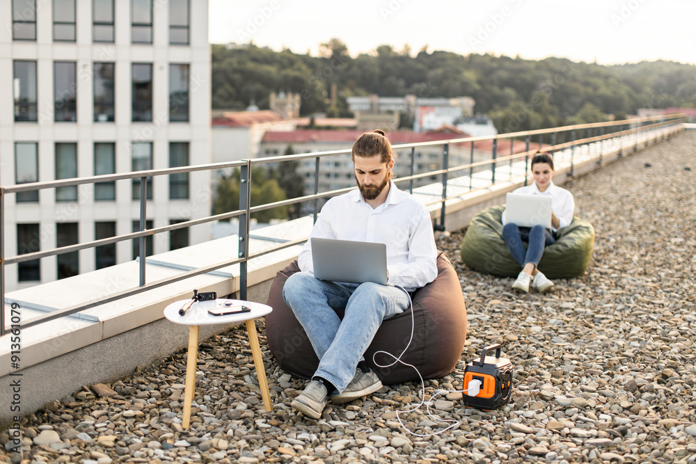 Business man sitting on bean bag chair connects charging cable to portable power station on rooftop of house. Relaxed outdoor work setting with laptops. Business man sitting on bean bag chair connects charging cable to portable power station on rooftop of house. Relaxed outdoor work setting with laptops.