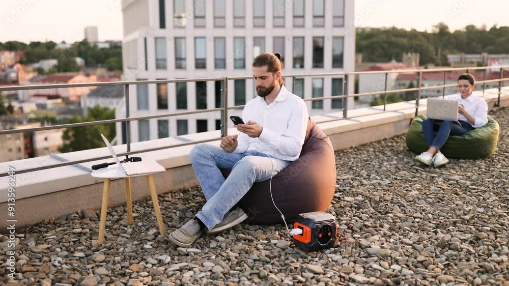 Businessman sitting on beanbag chair and plugs phone charging cable into portable power station on roof of house. Woman working on laptop in background. Outdoor office setup, remote work concept. Businessman sitting on beanbag chair and plugs phone charging cable into portable power station on roof of house. Woman working on laptop in background. Outdoor office setup, remote work concept.