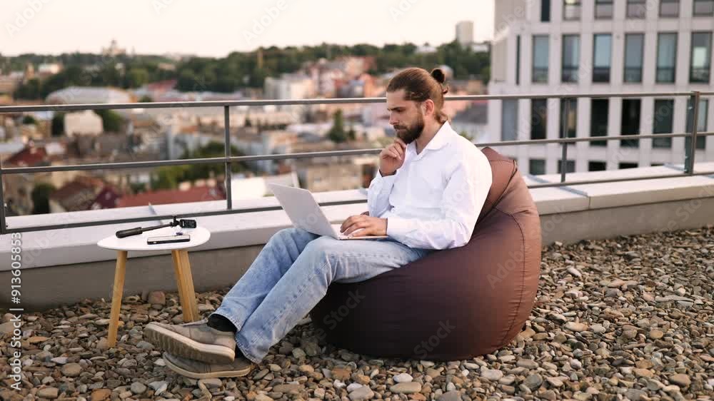 Man working on laptop sitting on beanbag outdoors on rooftop with cityscape in background. Concept of remote work, freelancing, and urban lifestyle. Man working on laptop sitting on beanbag outdoors on rooftop with cityscape in background. Concept of remote work, freelancing, and urban lifestyle.