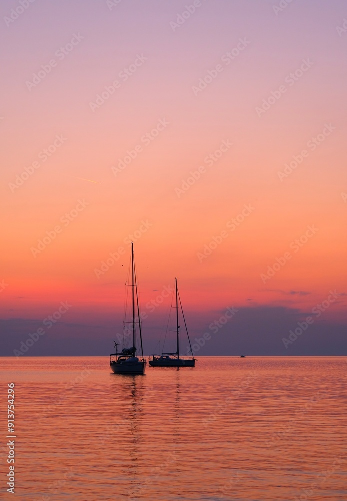 Two sailboats on a calm sea during a vibrant purple and orange sunset. The tranquil water reflects the stunning colors of the sky, creating a serene and picturesque scene. Nautical and sunset themes. Two sailboats on a calm sea during a vibrant purple and orange sunset. The tranquil water reflects the stunning colors of the sky, creating a serene and picturesque scene. Nautical and sunset themes.