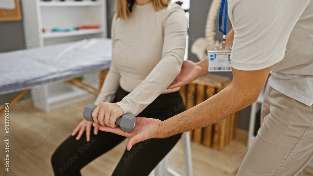 Obraz premium A woman in physiotherapy exercises her arm with a dumbbell assisted by a male therapist in a rehab center.